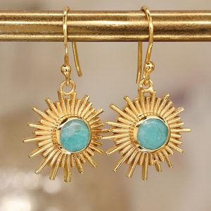 Könnte beinhalten: Ein Paar goldfarbene Ohrhänger mit einem Sonnenstrahlen-Design, bei dem strahlenförmige Metallstäbe einen runden, hellblauen Stein in der Mitte umgeben. Die Ohrringe hängen an einfachen, gebogenen Ohrhaken und haben eine glänzende Oberfläche.