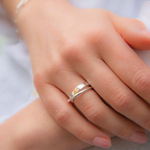 May include: A silver ring with a thin band and a thicker band that wraps around it. The thicker band has a subtle texture and a gold accent.