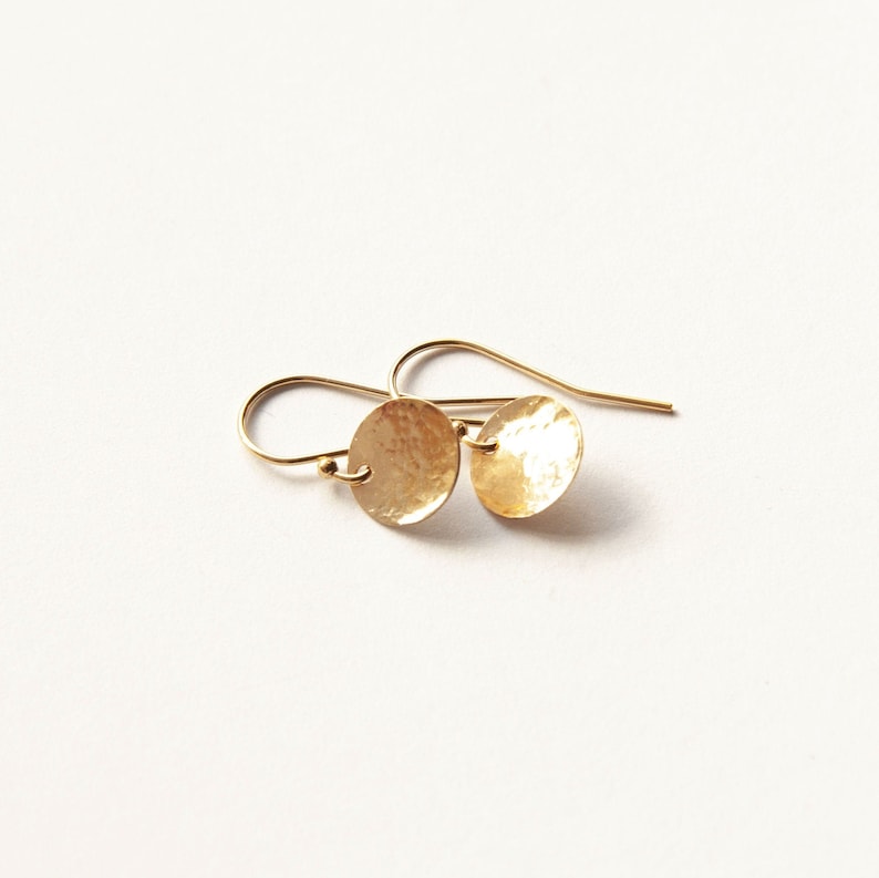 May include: A pair of gold-toned drop earrings featuring small, circular, hammered metal discs suspended from curved ear wires. The surface of each disc has a textured, reflective finish that catches the light, creating a bright, metallic appearance.
