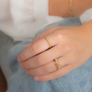 May include: Two gold rings on a hand. The top ring is a delicate chain with small flower-shaped links. The bottom ring is wider and features a row of colorful gemstones.