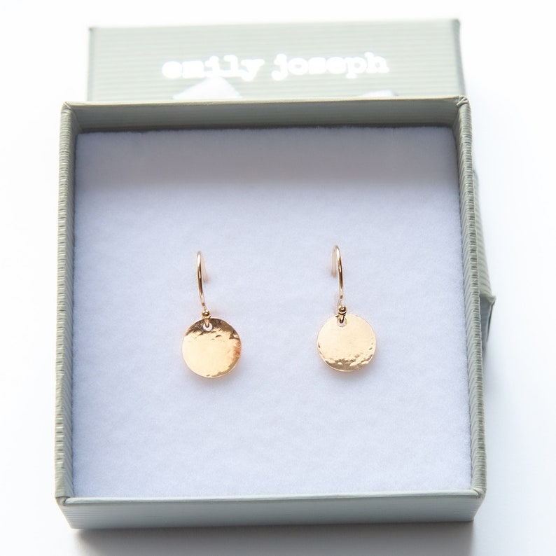 May include: A pair of rose gold-colored drop earrings featuring small, circular, hammered-texture discs suspended from curved ear wires. The earrings are presented inside a light gray gift box with the text emily joseph printed on the lid.