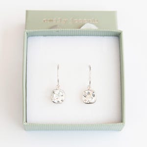 May include: A pair of silver dangle earrings with square-shaped clear crystal stones. The earrings are in a small green box with the text "emily jocelyn" on the top.