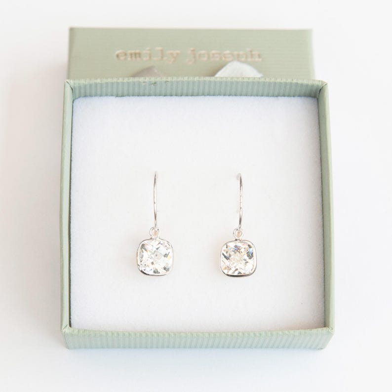 May include: A pair of silver dangle earrings with square-shaped clear crystal stones. The earrings are in a small green box with the text "emily jocelyn" on the top.