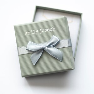 May include: A square, sage green jewelry box with a ribbed texture and a silver ribbon bow on top. The lid features the white text emily joseph. The box is slightly open, revealing a white interior and a thin gold chain necklace inside.