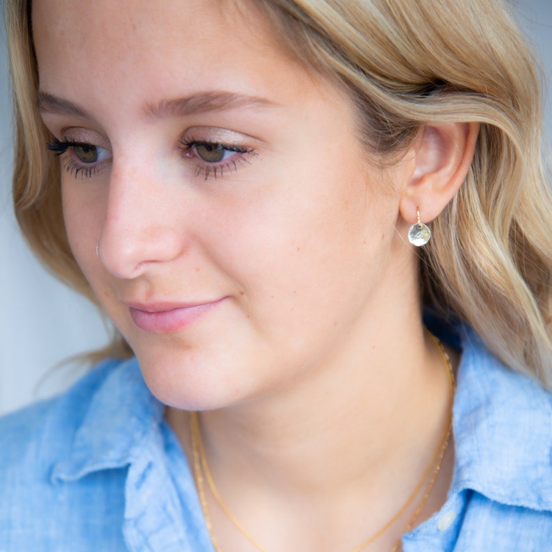 May include: A small, circular gold earring featuring a textured, hammered surface. The earring is designed as a delicate drop charm suspended from a thin gold hoop, creating a subtle and reflective metallic finish that catches the light.