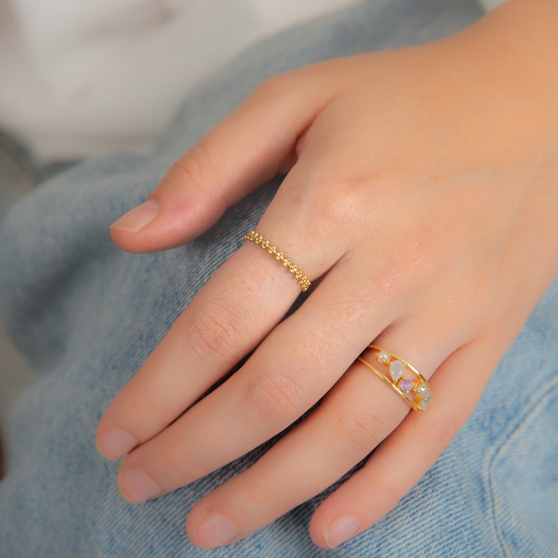 May include: Two gold rings on a hand. The top ring is a delicate chain with small beads. The bottom ring has a thin band with small stones in various colors.