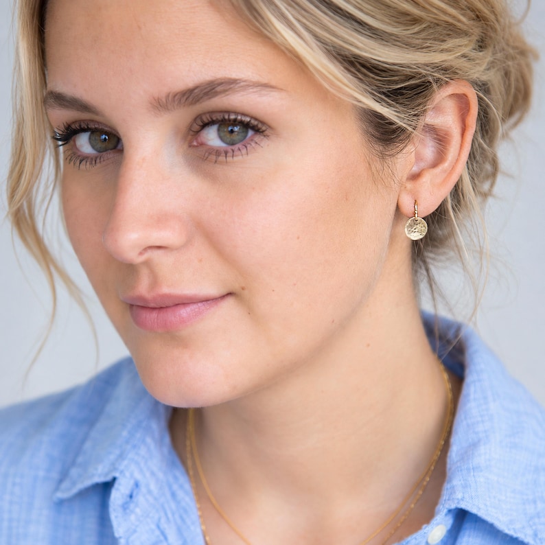 May include: A small, circular gold charm earring featuring a textured, hammered surface. The round pendant hangs from a thin, delicate gold hoop that secures through the earlobe. The metal has a warm, reflective finish that catches the light.