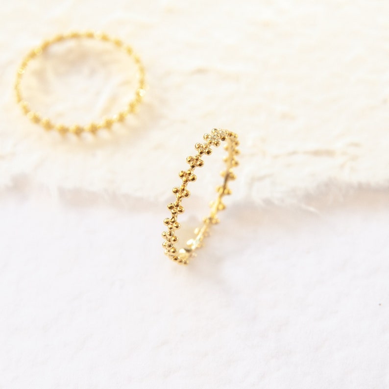 May include: Two gold rings with a delicate beaded design. The rings are on a white textured surface.