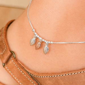 Peut inclure: Une chevillère en chaîne maille carrée argentée ornée de petites perles argentées et de trois breloques en forme de feuille. La feuille centrale est de couleur or rose, tandis que les deux feuilles extérieures sont en argent.