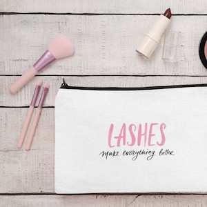 May include: White makeup bag with the text "LASHES make everything better" in pink and black. The bag is surrounded by makeup brushes, lipstick, a compact, and a makeup sponge on a white wooden surface.