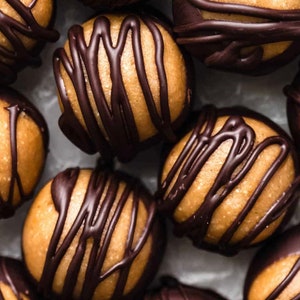 May include: Close-up of several chocolate-covered peanut butter balls. The balls are arranged in a cluster on a white surface. Each ball is covered in a thin layer of dark chocolate with drizzled lines of chocolate on top.