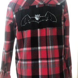 May include: Red and black plaid flannel shirt with a black patch on the back featuring a white embroidered bat.