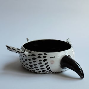 May include: A white ceramic bowl with a black and white spotted pattern and a black beak. The bowl has a black interior and a whimsical design.