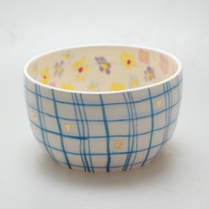 May include: A white ceramic bowl with a blue and white plaid pattern and gold accents. The bowl is decorated with small yellow, pink, and purple flowers.