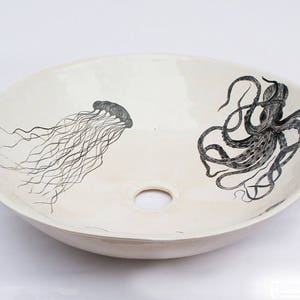 May include: Oval, off-white ceramic sink with a central drain hole. The sink features black illustrations of a jellyfish and an octopus. The jellyfish is on the left, and the octopus is on the right.