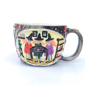 May include: A ceramic mug with a dark gray exterior and a whimsical design. The mug features a scene of three witches around a table, with a yellow background and decorative elements like mushrooms and stars. The handle is gray.