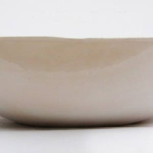 May include: A large, off-white ceramic bowl with a smooth, glossy finish. The bowl has a wide, shallow shape with gently curved sides and a slightly irregular rim. The interior appears to be a lighter shade of off-white. The bowl is likely used for serving or display.
