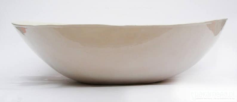 May include: A large, off-white ceramic bowl with a smooth, glossy finish. The bowl has a wide, shallow shape with gently curved sides and a slightly irregular rim. The interior appears to be a lighter shade of off-white. The bowl is likely used for serving or display.