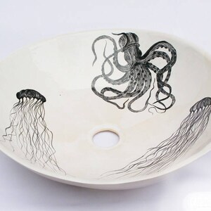 May include: Oval, off-white ceramic sink with a black octopus and jellyfish design. The octopus is centered, with two jellyfish on either side. The sink has a central drain hole, and the artwork is hand-drawn.