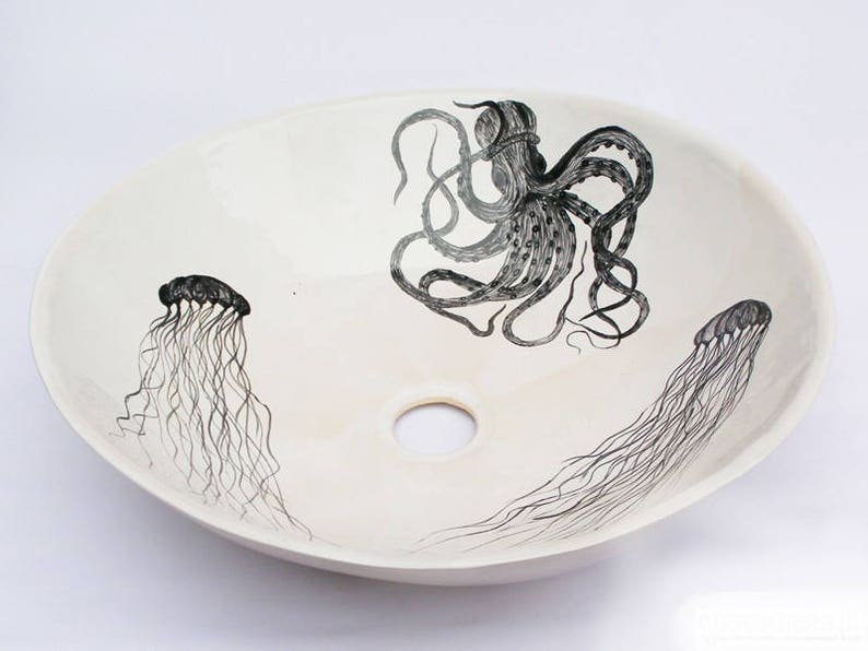 May include: Oval, off-white ceramic sink with a black octopus and jellyfish design. The octopus is centered, with two jellyfish on either side. The sink has a central drain hole, and the artwork is hand-drawn.
