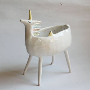 Può includere: Vaso da fiori in ceramica bianca a forma di unicorno con una criniera arcobaleno e un interno macchiato. Il vaso ha quattro gambe e si trova su una superficie bianca.