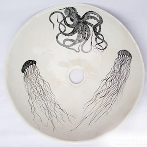 May include: A round, off-white ceramic sink basin featuring a hand-drawn octopus and two jellyfish in black ink. The octopus is at the top, and the jellyfish are on either side of the drain hole.