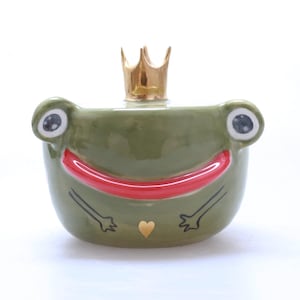 May include: A green ceramic frog figurine with a gold crown, red smile, and gold heart. The frog has black eyes and arms.
