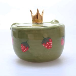 May include: A green ceramic jar with a gold crown lid and a strawberry design. The jar has a round shape and a smooth surface.