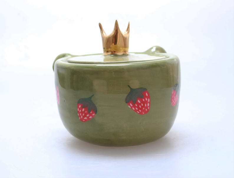 May include: A green ceramic jar with a gold crown lid and a strawberry design. The jar has a round shape and a smooth surface.