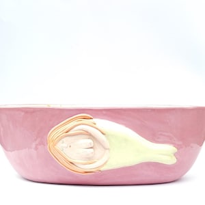 Op de afbeelding: Een roze keramische kom met een glanzende afwerking. De kom heeft een gebeeldhouwde figuur van een persoon met lang haar en een lichtgroen kledingstuk. De figuur ligt met gesloten ogen. De kom is ovaal van vorm.