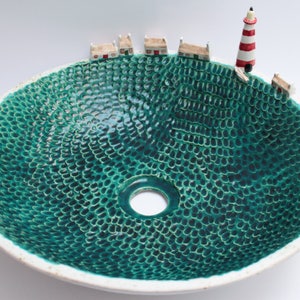 May include: A ceramic sink basin with a textured teal interior and a white rim. Miniature houses and a red and white striped lighthouse are arranged around the edge, creating a coastal village scene.
