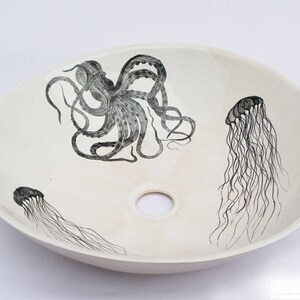 May include: Oval, off-white ceramic sink with a central drain hole. The sink features black illustrations of an octopus and two jellyfish. The octopus is centrally located, while the jellyfish are positioned on either side. The design is nautical themed.