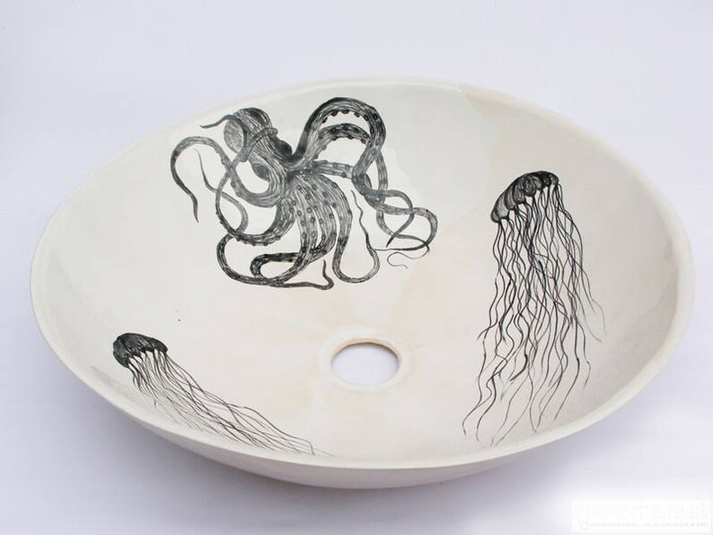 May include: Oval, off-white ceramic sink with a central drain hole. The sink features black illustrations of an octopus and two jellyfish. The octopus is centrally located, while the jellyfish are positioned on either side. The design is nautical themed.