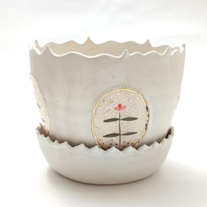 May include: A white ceramic planter with a matching saucer. The planter features a scalloped rim and is decorated with gold-rimmed oval designs with a pink flower and green stem. The planter is designed for indoor or outdoor use.
