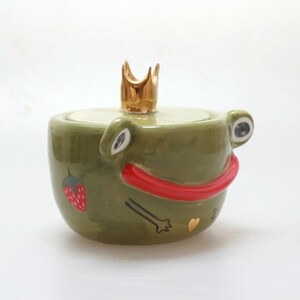 May include: Green ceramic frog-shaped jar with a gold crown on top. The frog has red lips, a strawberry, and a gold heart on its body.