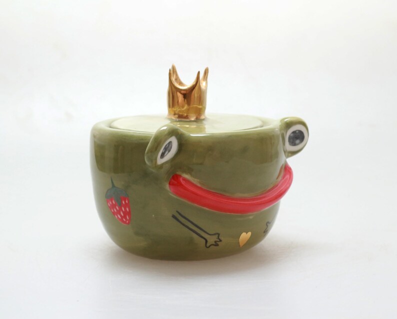 May include: Green ceramic frog-shaped jar with a gold crown on top. The frog has red lips, a strawberry, and a gold heart on its body.