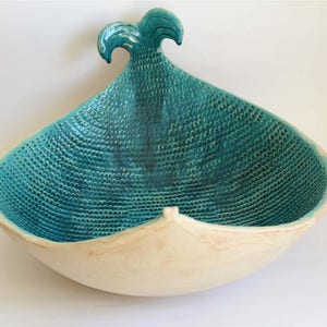 May include: A teal blue ceramic bowl with a textured surface and a wave-shaped handle. The bowl is white on the outside and has a unique, organic shape.