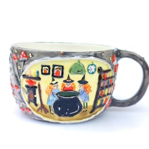 May include: A ceramic mug with a whimsical witch-themed design. The mug features a gray exterior with red mushroom accents and a yellow interior scene depicting three witches around a cauldron. A broom, bookshelf, and clock are also visible.