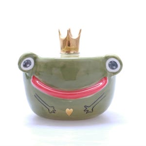 May include: A green ceramic frog figurine with a gold crown on its head. The frog has a red smile and a gold heart on its chest.