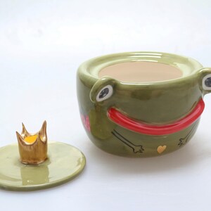 May include: A green ceramic frog-shaped container with a red mouth and a gold crown lid. The container has a heart and arrow design on the side.