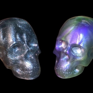 May include: Two iridescent skull sculptures. One is silver and the other is a mix of purple, green, and blue. The skulls are side-by-side against a black background. The skulls are detailed and appear to be made of resin.