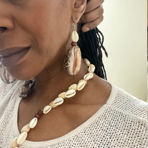 May include: A seashell jewelry set featuring a necklace and earrings. The necklace is made of small, white seashells and interspersed with brown beads. The earrings have a large, oval-shaped shell with a smaller white shell and a brown bead above.