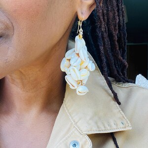 Peut inclure: Boucles d'oreilles composées de grappes de petits coquillages crème. Les coquillages sont assemblés avec du fil doré, créant un design pendant, de style chandelier. Les boucles d'oreilles sont suspendues à des crochets d'oreilles dorés.