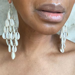 Peut inclure: Gros plan d'une personne portant des boucles d'oreilles pendantes en coquillages blancs. Les boucles d'oreilles sont attachées à une chaîne argentée et sont disposées en forme de lustre. Les lèvres de la personne sont visibles sur l'image.