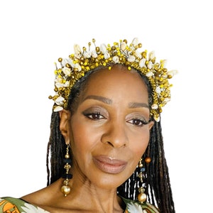 Può includere: Una donna indossa una corona di perline dorate e bianche e orecchini abbinati. La corona presenta un design a forma di alone con perline bianche a forma di conchiglia e accenti dorati. Gli orecchini hanno elementi dorati, neri e marroni.