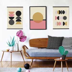 May include: Three framed abstract geometric prints with black, white, gray, pink, and yellow shapes on a cream background. The prints are hanging on a white wall above a mid-century modern sofa with gray cushions and a green cactus on a wooden side table.