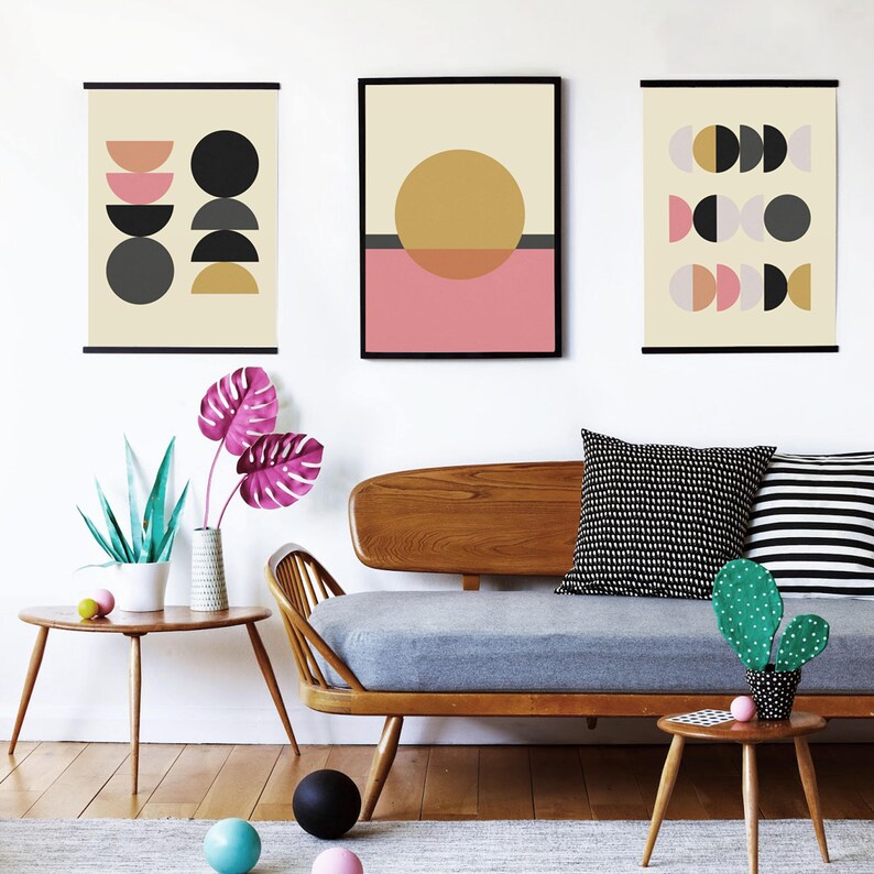 May include: Three framed abstract geometric prints with black, white, gray, pink, and yellow shapes on a cream background. The prints are hanging on a white wall above a mid-century modern sofa with gray cushions and a green cactus on a wooden side table.