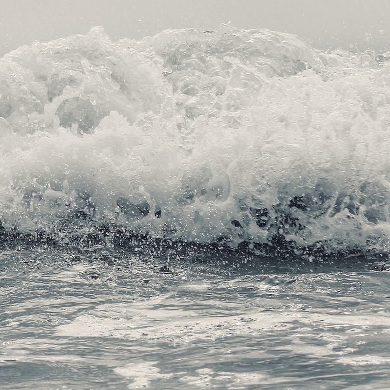 May include: A close-up of a wave crashing on the shore, creating a white foamy crest. The water is a light blue-gray color.