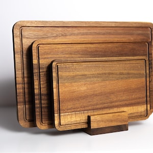 May include: A set of three rectangular wooden cutting boards of varying sizes, each featuring a dark brown wood grain and a carved juice groove along the perimeter. The boards are nested upright in a matching wooden stand for storage and display.