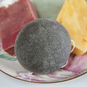 Puede incluir: Una esponja facial de carbón vegetal negra en un plato blanco con diseños florales rosas y morados. Dos barras de jabón, una rosa y otra amarilla, también están en el plato.